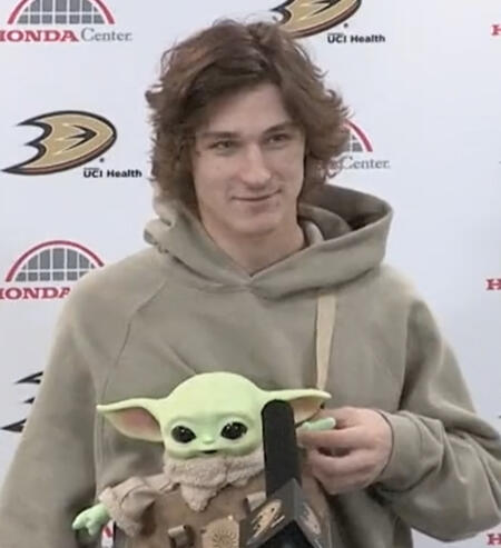 trevor zegras, holding up the hand of a plush yoda strapped to his chest in a wave