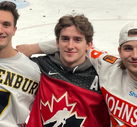 mackie samoskevich smiles lightly at the camera, his red jersey contrasting with the white background nicely. he has two unseen teammates arms wrapped around his shoulders